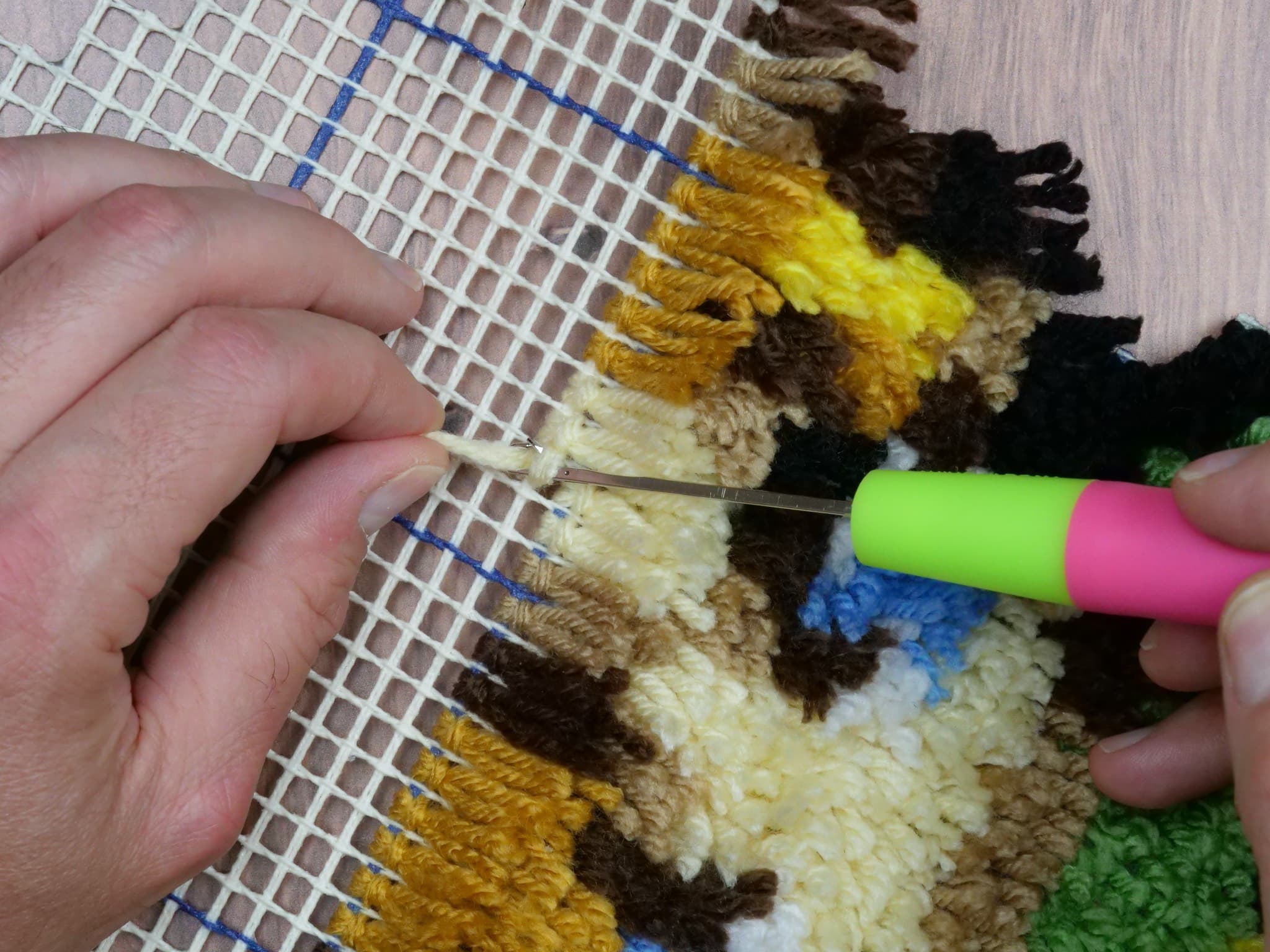 Hands working yarn through a latch hook canvas.