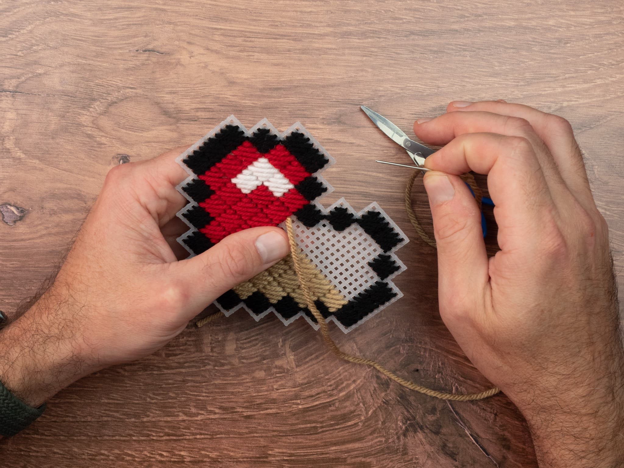 Hands stitching yarn through a plastic canvas pixel design.